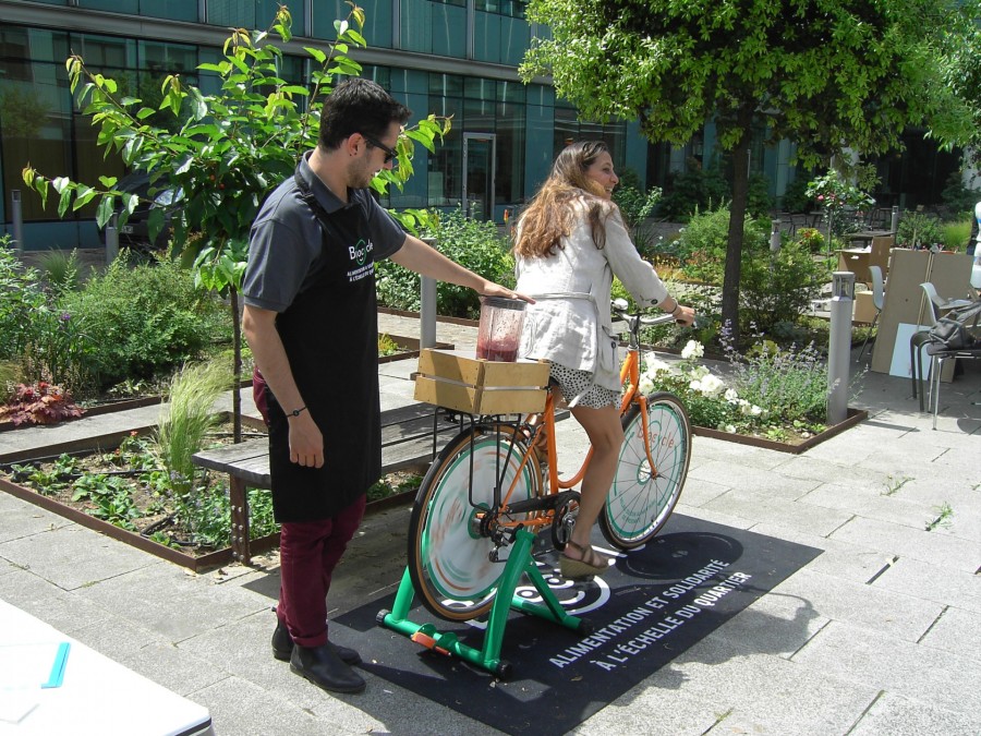 Une femme pédale sur le vélomixeur, un bénévole Biocycle derrière elle